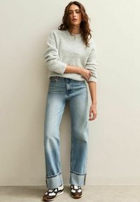 Vrouw in een lichtgrijs fuzzy sweater, met breedomgeslagen lichtblauwe jeans en bruin-witte sneakers, staande tegen een beige muur.