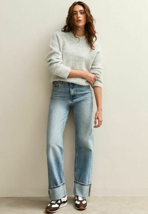 Vrouw in een lichtgrijs fuzzy sweater, met breedomgeslagen lichtblauwe jeans en bruin-witte sneakers, staande tegen een beige muur.