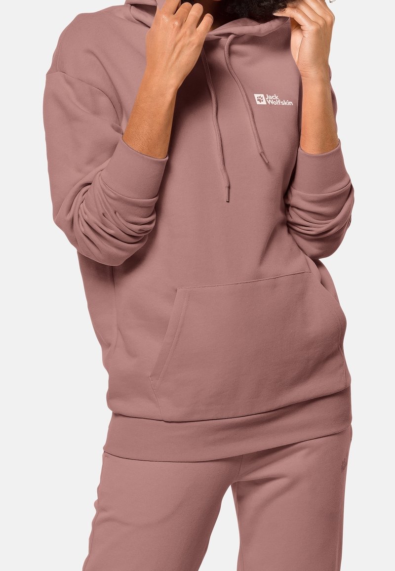 Persona que lleva una sudadera con capucha Jack Wolfskin color malva y pantalones a juego, ajustando los cordones de la capucha con las manos sobre un fondo liso.