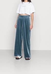 Žena ve bílem krátkém tričku s nápisem „MINIMALIST“, modrých sametových kalhotách širokého střihu a bílých sandálech s tlustou černou podrážkou.