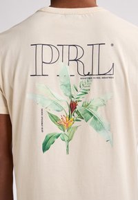 Camiseta beige con cuello redondo que presenta una gran estampado botánico de hojas verdes y flores naranjas, etiquetada "PIRL" en letras negras en negrita.