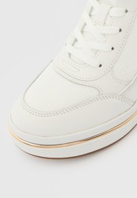 Zapatilla blanca de malla texturizada y cuero liso, con cordones planos, una franja decorativa dorada y un diseño de puntera redondeada.