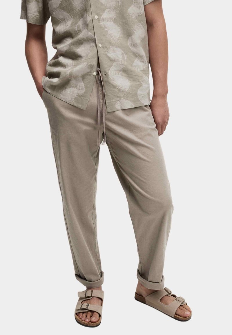 Personne portant une chemise beige à manches courtes à motifs, un pantalon beige ample avec cordon, et des sandales beiges à double bride, debout avec une main dans la poche.