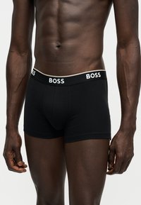 Mužský trup a horné končatiny v čiernych boxerkách BOSS s označeným pásom, stojaci pred jednoliatym svetlým pozadím.