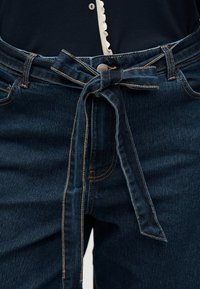 Σκούρο μπλε τζιν παντελόνι με υφασμάτινη ζώνη δεμένη σε φιόγκο στη μέση, φορεμένο με σκούρο τοπ που έχει ένα μικρό λευκό κουμπί και διακόσμηση με βολάν.