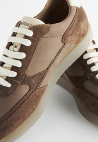 Brune ruskind- og lædersneakers har cremede snørebånd, kontrasterende teksturer og en klar gummisål med synlige syninger.