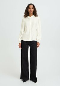 Blusa de mangas largas color crema con cuello, que presenta un cierre de botones en la parte delantera y una tela texturizada. Combinada con pantalones de pierna ancha negros.