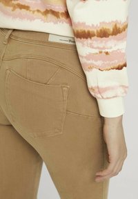 Vaqueros de mezclilla beige con un diseño ajustado, con un único bolsillo trasero y combinados con una sudadera de mangas con un patrón en rosa y crema.