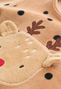 Beige Fleece-Stoff mit einem Rentierdesign, das braune Hörner, eine rote Nase und weiße gestickte Details aufweist. Schwarze Polka-Dots als Akzente.