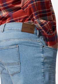 Jeans de acabado en denim azul claro, con una etiqueta de cuero marrón que dice "JOHN F. GEE" en la parte de atrás, combinados con una camisa de cuadros roja y azul.