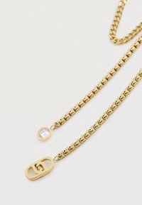 Χρυσή αλυσίδα κολιέ με δύο κλωστές· η μία έχει ένα στρογγυλό κρύσταλλο ως κρεμαστό στοιχείο, ενώ η άλλη διαθέτει ένα ορθογώνιο γούρι με εγ engraved λόγω του λογότυπου.