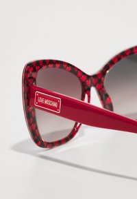 Red sunglasses with a heart-patterned frame, large cat-eye shape, gradient lenses, and a gold "Love Moschino" logo on the arm.