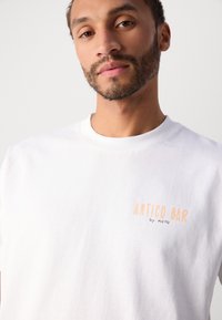 Witte katoen T-shirt met ronde hals, voorzien van "ANTICO BAR by MOPO" in oranje tekst op de borst. Glad textuur, casual pasvorm.