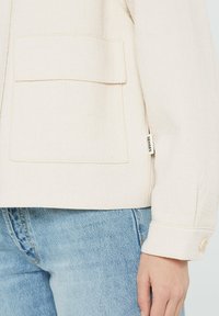 Cream textured jacket with a flap pocket and buttoned cuff worn with light blue denim jeans, showing a "SENSES" fabric tag on the side seam.