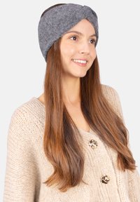 Un bandeau en tissu gris texturé avec un design frontal torsadé, porté avec un pull en maille épaisse crème comportant de boutons ronds brillants.