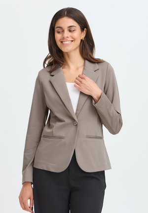Jonge vrouw glimlacht, draagt een taupekleurige blazer over een wit topje en zwarte broek, met haar rechterhand die de revers van het blazer aanraakt.