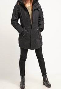 Sort parka med hætte i pels, lynlås og trykknaplukning, to sidelommer og justerbare manchetter, parret med sorte slim-fit bukser og ankelstøvler.