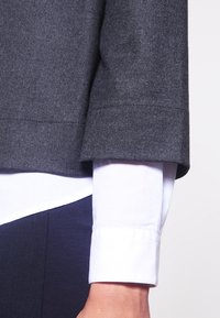Top in misto lana grigio scuro con design cropped, caratterizzato da un risvolto sopra una camicia bianca. Il tessuto ha una consistenza liscia.