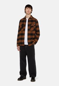 Brun og sort ternet flannelskjorte med to brystlommer, båret åben over en hvid t-shirt, parret med sorte vide bukser og sorte sko.