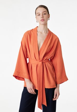 Jonge vrouw met een oranje kimono-jas met brede mouwen en een ceintuur, en zwarte broek, staand tegen een witte achtergrond.