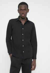 Camisa negra abotonada de tela texturizada con cuello, botones blancos y mangas largas, combinada con pantalones oscuros.