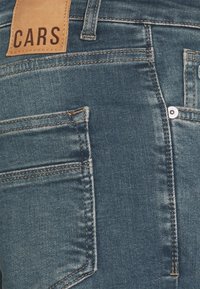 Dark blue denim jeans featuring a leather patch labeled "CARS" and accented with contrasting stitching and a back pocket detail.