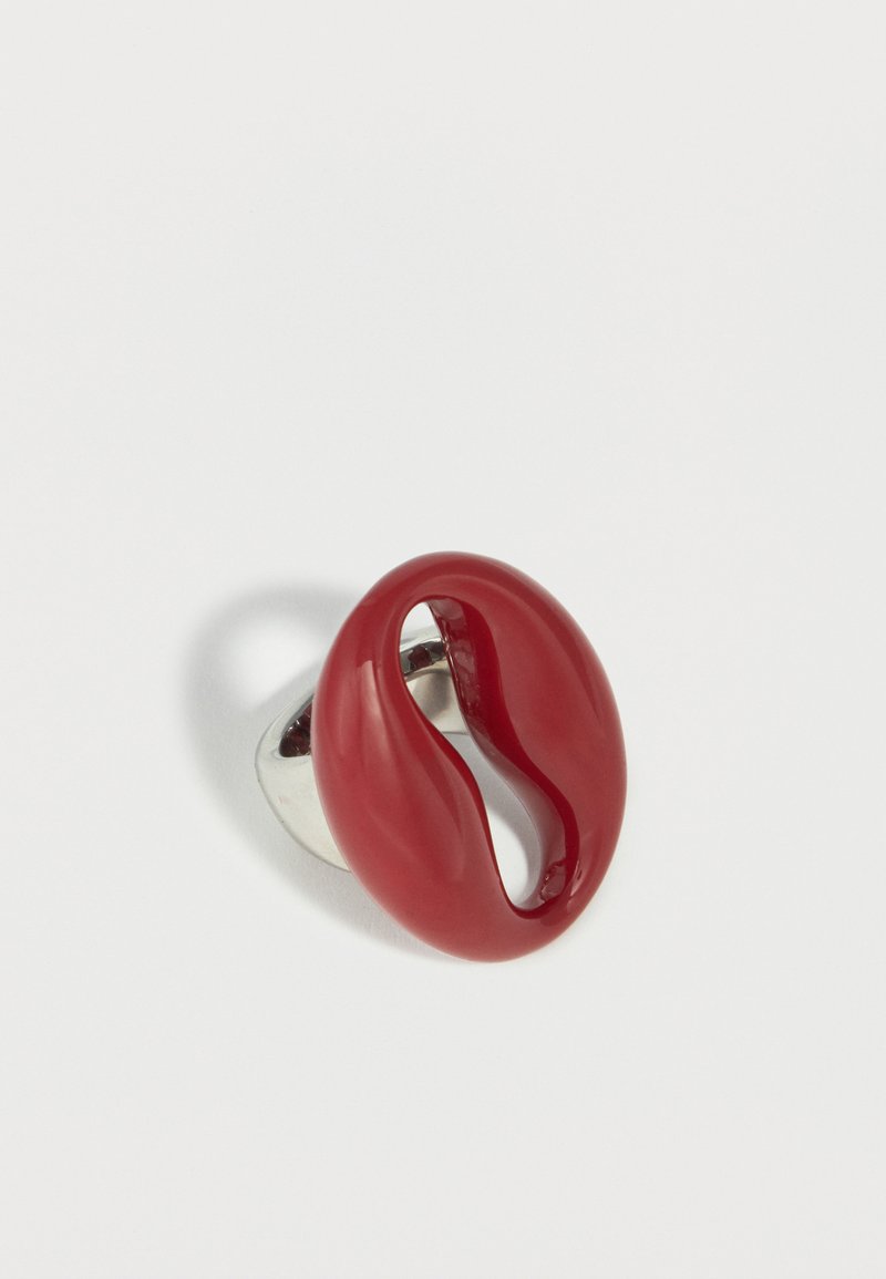Bague moderne rouge brillante de forme abstraite sur un anneau réglable en argent posée sur une surface blanche.