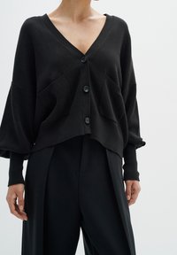 Černý kardigan na knoflíky z měkké pletené látky. Má hluboký výstřih do V, dvě přední kapsy a volné, nadměrně velké rukávy.