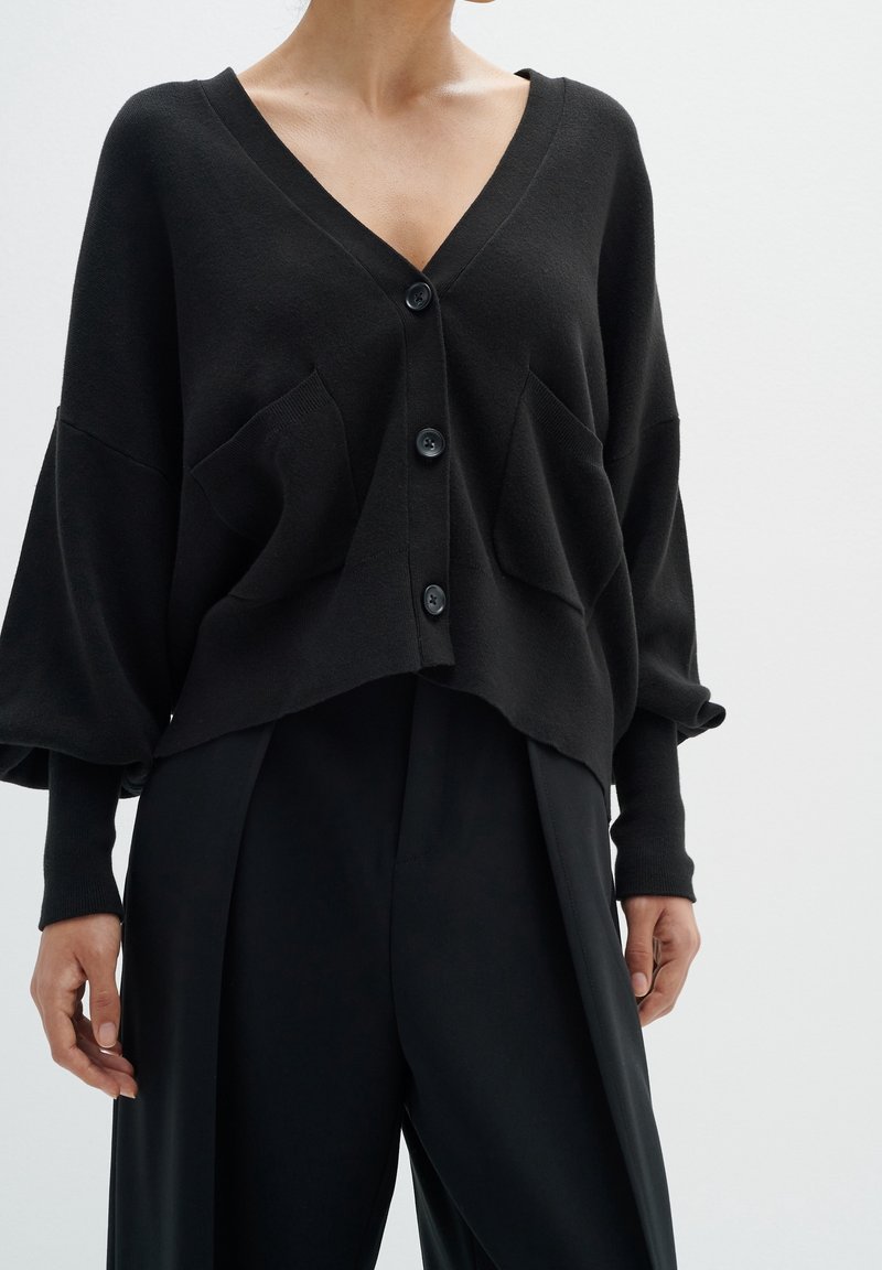 Černý kardigan na knoflíky z měkké pletené látky. Má hluboký výstřih do V, dvě přední kapsy a volné, nadměrně velké rukávy.