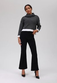 Svart og hvit stripete genser med høy hals og ribbestruktur i kanten, kombinert med svarte flagrende bukser og svarte høye hæler, posert mot en nøytral bakgrunn.