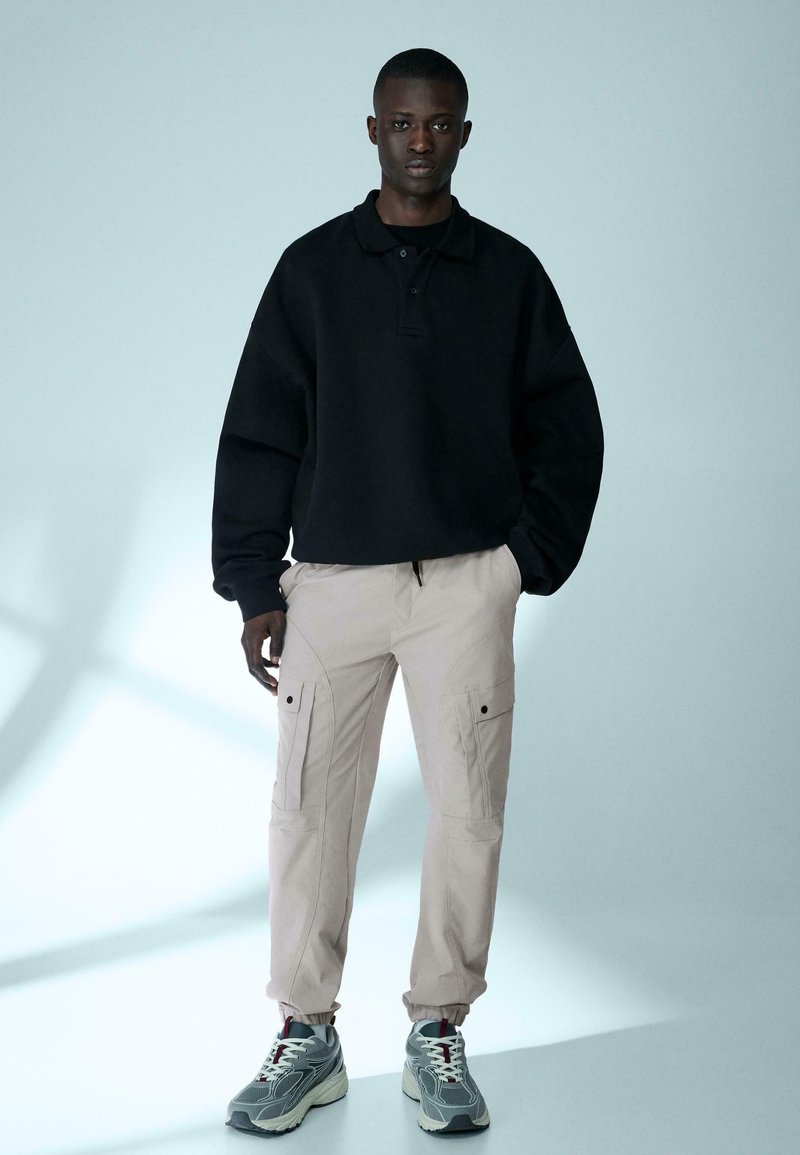 Un jeune homme se tient debout, portant un polo noir à manches longues, un pantalon cargo beige et des baskets grises, sur un fond bleu clair uni.
