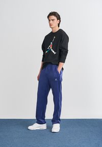 Μαύρο φούτερ με μακριά μανίκια και πολύχρωμο γραφικό, συνδυασμένο με μπλε φόρμες που έχουν λευκές ρίγες στο πλάι και λογότυπο της Jordan. Λευκά αθλητικά παπούτσια.