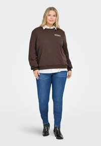 Žena v hnědé mikině s nápisem "ESPRESSO YOURSELF", bílé košilce, modrých džínách a černých kotníkových botách, stojící na jednobarevném pozadí.