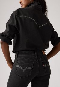 Sort denimjakke med en robust tekstur og oprullede ærmer, der har dekorative sølvnitter på ryggen og matchende sorte jeans.