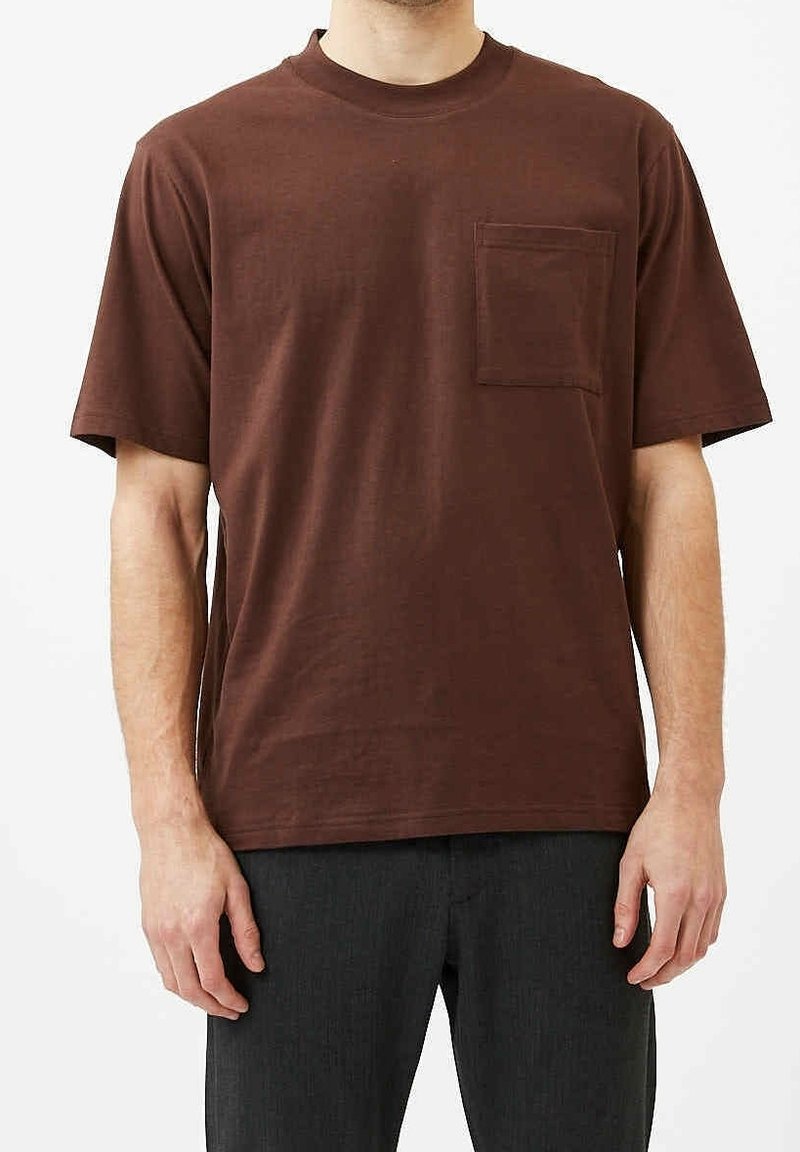 Homme portant un T-shirt marron uni à manches courtes avec une poche sur la poitrine et un pantalon foncé, debout devant un fond blanc.