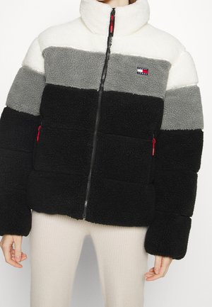 Giacca in pile con blocchi di colore orizzontali in bianco, grigio e nero, chiusura frontale con zip, colletto alto e toppa con logo sul petto. Dettagli rossi sulla zip.