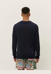 Granatowy sweter z prążkowanej struktury z długimi rękawami i okrągłym dekoltem, z teksturowanym wzorem, w połączeniu z szortami w wzory. Pokazano widok z tyłu.