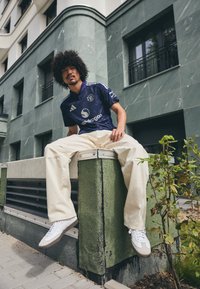 Fialový dres Manchester United s bílými akcenty, béžové oversize kalhoty a bílé tenisky s šedými detaily, sedící na zelené texturované lavičce.