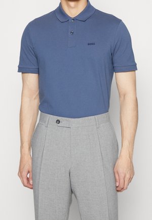 Man in een blauwe korte mouwen polo die in lichtgrijze pantalon is gestoken, staande tegen een effen achtergrond.