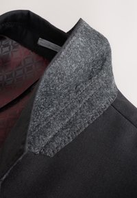 Col d'une veste noire avec un détail en tissu gris texturé à l'intérieur et une doublure rouge à motifs visible sous le col.
