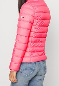 Růžová prošívaná bunda typu puffer s límcem stojačkem a logem na levém rukávu. Hladký, lesklý materiál, vyprofilovaný pas.