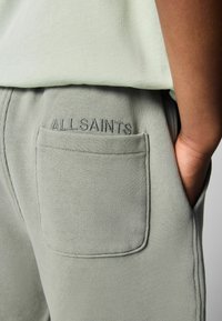 Světle zelené tepláky s měkkou texturou, s zadní kapsou a vyraženým nápisem "ALLSAINTS" na pasu.