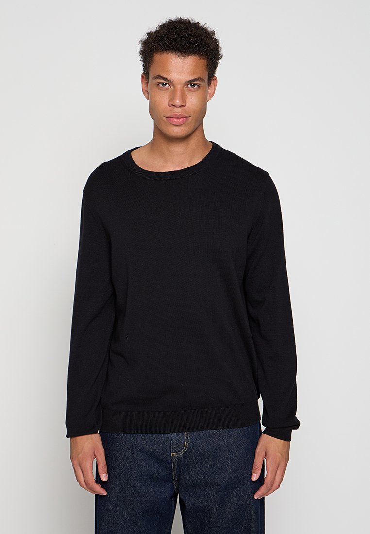 Joven con pelo corto y rizado, vestido con un suéter negro de manga larga y unos pantalones vaqueros azul oscuro, de pie frente a un fondo claro.