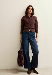 Brązowy sweter z dzianiny z guzikami, ciemnoniebieskie szerokie spodnie z wysokim stanem, bordowa błyszcząca torba tote oraz brązowe skórzane botki z wzorem w panterkę.