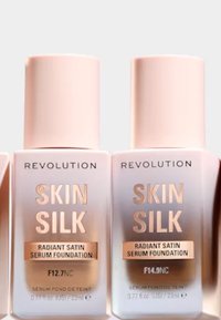 To Revolusjon Skin Silk Radiant Satin Serum Foundation flasker. Lys rosa plastbeholdere med gullaccenter, etiketter som viser skyggenumrene F12.7NC, F14.9NC.