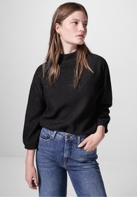 Maglione nero lavorato a maglia con maniche a sbuffo lunghe e colletto alto, caratterizzato da un motivo testurizzato. Abbinato a jeans in denim blu.