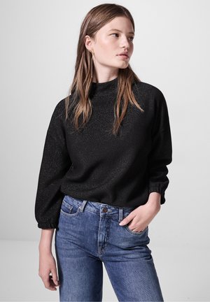 Schwarzer Strickpullover mit langen Puffärmeln und hohem Kragen, der ein strukturiertes Muster aufweist. Kombiniert mit blauen Jeans.