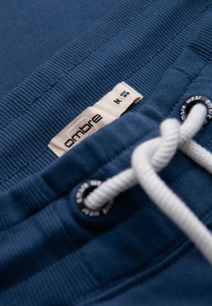 Zbliżenie na pas od niebieskich spodni dresowych z białym sznurkiem, metalowymi oczkami i beżową metką z rozmiarem "ombre M".