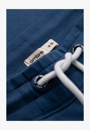 Primo piano della vita di pantaloni sportivi blu con coulisse bianca, occhielli in metallo e un'etichetta beige con la taglia "ombre M."