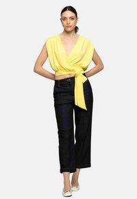 Top giallo avvolgente con una cintura in vita, abbinato a jeans di jeans scuri. Il top presenta un profondo scollo a V e maniche corte.